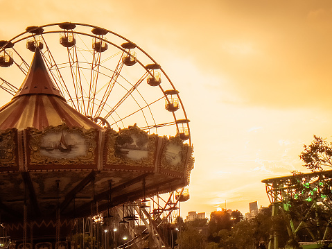 Petaling Jaya, Malaysia – April 04, 2023: Sun setting behind 2 fair ground structures which are the carrousel and ferris wheel, setting the mood to a night of neon lights and loud amusement sounds.