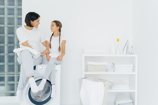Caring mother talks with daughter, pose on washing machine, surrounded with white linen, do washing together, being in laundry room at home. Happy adult housewife thanks adorable girl for help