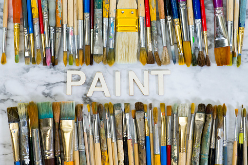 Artist's used paintbrushes in assorted colours and sizes lined up above and below the word paint spelled out in wooden letters on a white marble background.