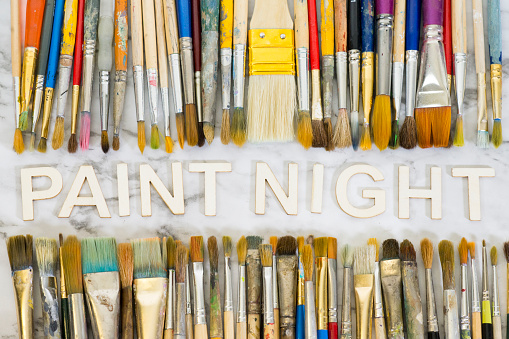 Artist's used paintbrushes in assorted colours and sizes lined up above and below the words paint night spelled out in wooden letters on a white marble background.
