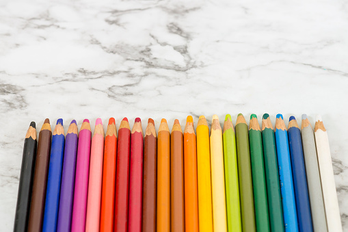 Wooden artists rainbow of drawing pencil crayons are laying in a straight line on a white marble background with copy space above them.