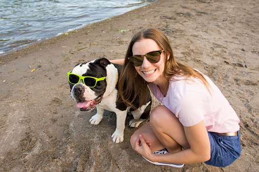 Happy young female pet owner smiling a toothy smile as she and her pet English bulldog are wearing sunglasses and posing for a portrait on the beach on a hot summer day.
