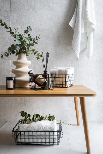 Concept of bathroom interior decor and cosmetics. Folded white towels in metal baskets, shower gel in dispenser bottle, incense sticks, ceramic vase with green eucalyptus branch on wooden table