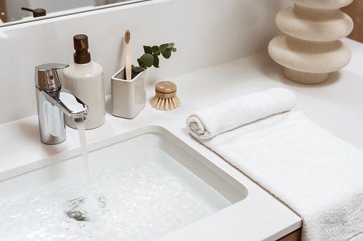 Hight angle view of white washbasin filled water, metal faucet, dispenser, toothbrush in a cup, bamboo brush, rolled towel and ceramic vase in a white furniture