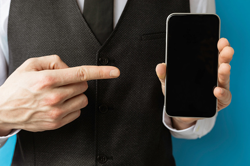 Finger of unrecognizable businessman is pointing at phone in front of blue background.