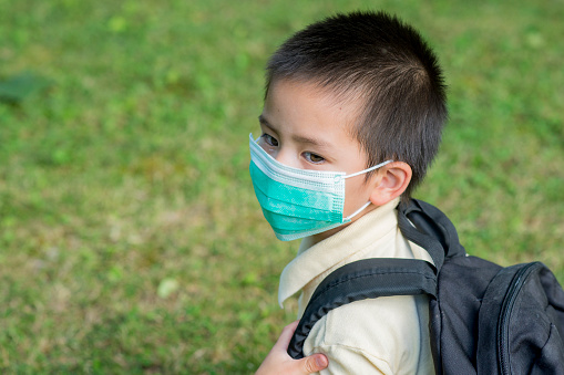 Cute young Malaysian Canadian boy wearing a medical mask to protect him from Covid-19 and school backpack looking back with a serious expression. He is ready to go back to school during the coronavirus pandemic.