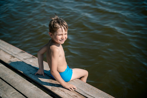 cute caucasian boy sitting on wooden pier diving into lake in countryside