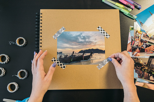 Image seen from above of a middle-aged woman's hands sticking a small piece of washi tape on the cover of a handmade kraft paper travel photo album.