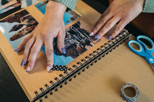 Close-up image of the hands of an unrecognizable middle-aged woman making sure that the piece of washi tape over a photo in her handmade kraft travel album is securely attached.