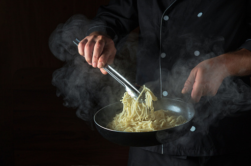 Cooking spaghetti in a frying pan in the hands of a chef. Space for advertising on a black background