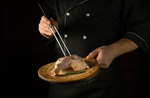 The chef prepares lunch with raw chicken leg on the cutting board of the restaurant kitchen. Delicious idea for grilling on black background.