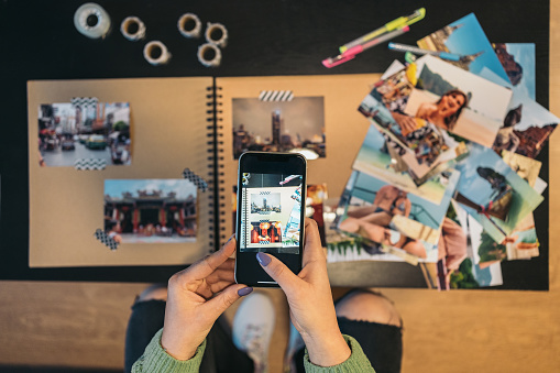 Close-up image seen from above of the hands of an unrecognizable middle-aged woman taking a photograph with her mobile phone of her handmade kraft travel album made with washi tape.