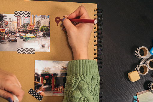 Image from above of an unrecognizable young woman's hand filling in a red location marker with a felt-tip pen on a handmade kraft album with travel photos and washi tape.