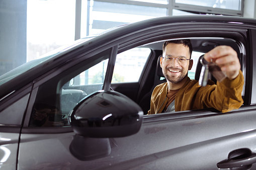 Happy man holding key for his new car in a showroom and looking a the camera