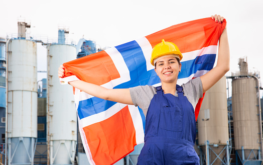 Positive young female worker in helmet waving national flag of Norway while standing in front of big tanks at refinery factory on sunny summer day