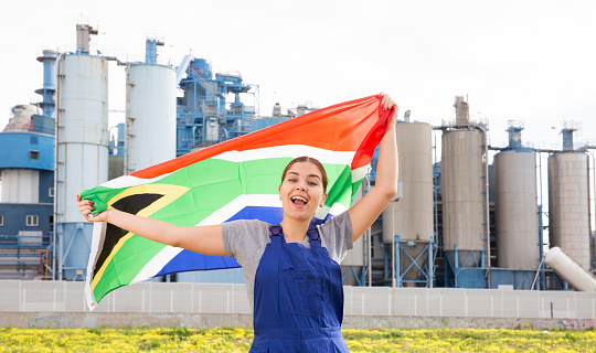 Smiling young woman in uniform posing with South Africa flag near factory