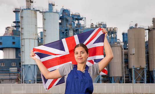 Against backdrop of factory located outside city,balanced girl in overalls of worker stands and holds flag of Great Britain in her hands.