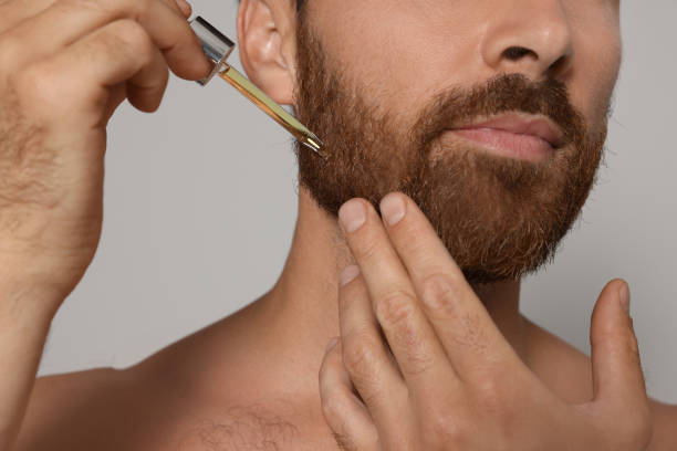 Man applying oil onto beard on grey background, closeup