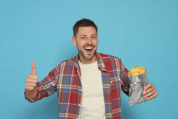 istock Handsome man with potato chips showing thumb up on light blue background 1483392398