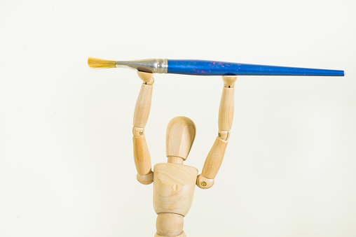 Artist's small wooden drawing model posing holding a blue wide paint brush high above its head on a blank white background.