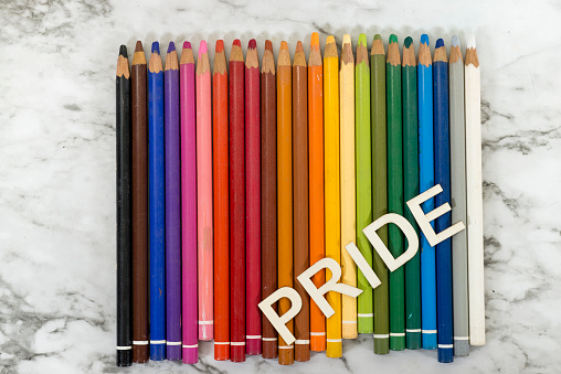 Wooden artists rainbow of drawing pencil crayons are laying in a straight line on a white marble background with copy space above them. The word pride is spelled diagonally across them in wooden letters to celebrate LGBT people.