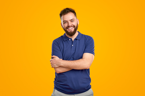 Happy young bearded guy in blue polo shirt smiling and looking at camera while standing against bright yellow background with crossed arms