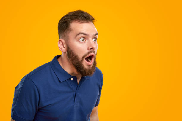 Astonished bearded man in blue t shirt looking away with opened mouth against yellow background