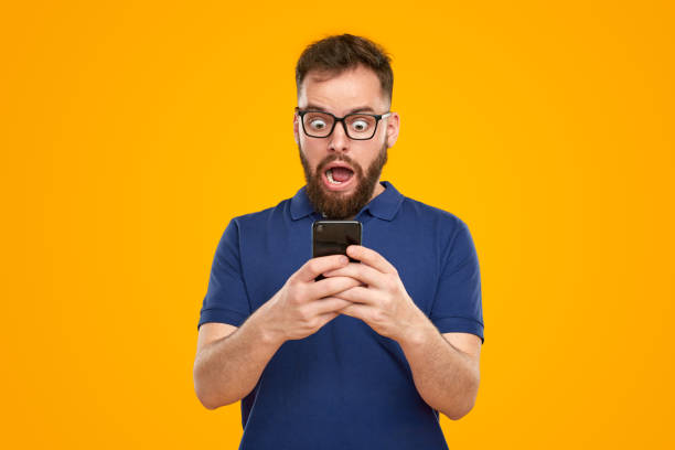 Surprised man reading news on smartphone Funny astonished bearded male in glasses reading shocking news on mobile phone, and looking at screen with eyes wide open on yellow background in studio stressed man funny stock pictures, royalty-free photos & images