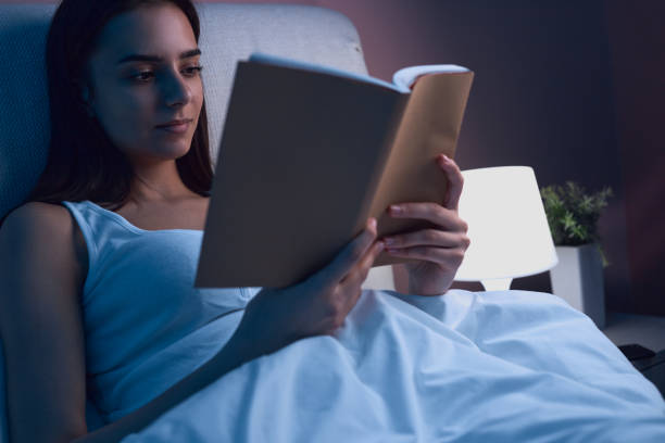 Woman reading book in bed before sleep Peaceful female lying in bed under soft blanket and reading interesting story in book while spending evening in bedroom before falling asleep low lighting stock pictures, royalty-free photos & images