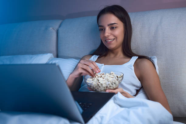 istock Smiling young ethnic lady lying on bed with popcorn bowl and watching laptop 1483417029