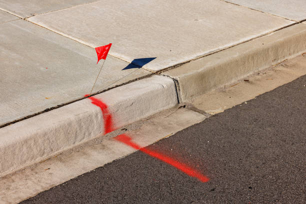 Small red flag label and spray painted marking line on pavement curb as identification of underground communications. Flag casts shadow on a ground