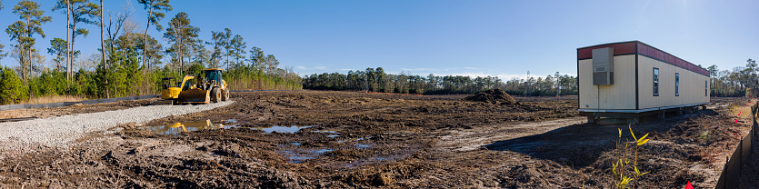 Clearing the way. Construction site with bulldozer and trailer for workers on dry dirty grass area surrounded by trees. Stitched panorama