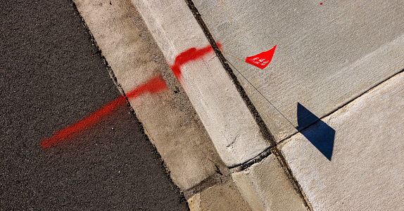Small red flag label and spray painted marking line on pavement curb as identification of underground communications. Flag casts shadow on a ground