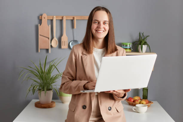 femme indépendante souriante heureuse aux cheveux bruns portant une veste beige debout à l’intérieur de la cuisine travaillant sur un ordinateur portable de la maison se préparant pour une réunion en ligne. - women computer home interior brown hair photos et images de collection