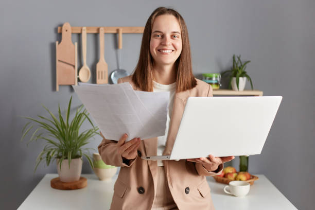 베이지색 재킷을 입은 갈색 머리의 행복한 여자는 원격 작업을 하는 서류를 들고 있는 노트북 컴퓨터를 사용하여 집 부엌 내부에 서 있다. - women computer home interior brown hair 뉴스 사진 이미지