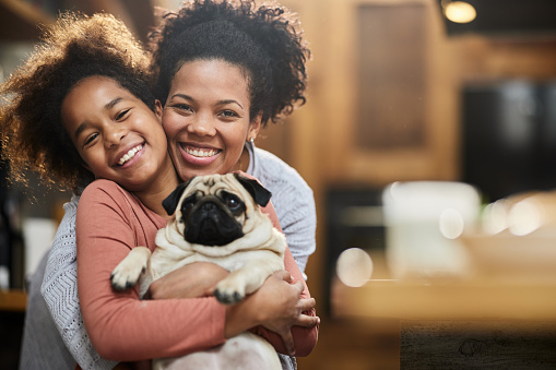 Portrait of happy African American mother and daughter enjoying while embracing their dog at home and looking at camera.
