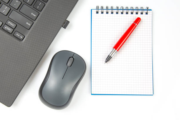 istock Computer wireless mouse, pen and notepad next to a laptop on a white table close-up. Items for office work 1483474428