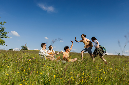 Group of happy friends relaxing in grass in a meadow while couple is coming to be with them. Copy space.