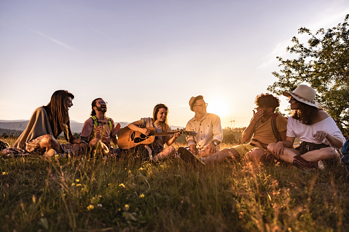 Group of young happy hikers enjoying in guitar music while resting on a hill at sunset. Copy space.