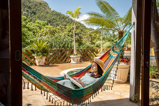 Beautiful young woman relaxing in a hammock and reading a book at vacation rental home during summer