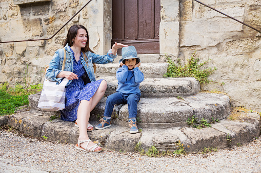 Mother and little handsome baby boy sitting on ancient stone stairs and playing outdoor with straw hat in old town