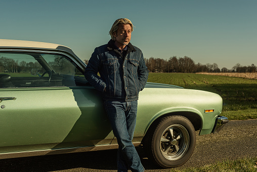 Man with blond hair in jeans stands on front side of a vintage american muscle car in sunny countryside.