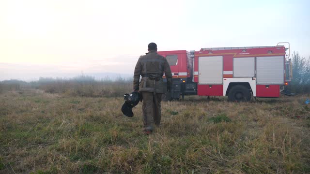 Male firefighter in uniform going on dry grass at countryside. Professional fireman walking after hard work to fire truck. Young fireguard with helmet in hand moving through uncultivated meadow