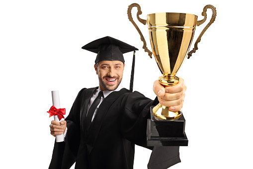Full length portrait of a happy young man wearing a graduation gown and holding a gold trophy cup isolated on white background