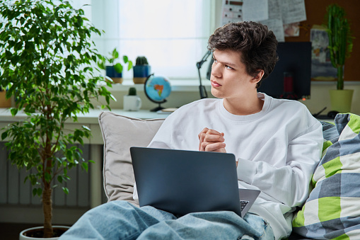 Teenage guy lying at home on couch student using laptop notebook textbook. Handsome male teenager college, high school student learning online remotely. Education, learning, technology, youth concept