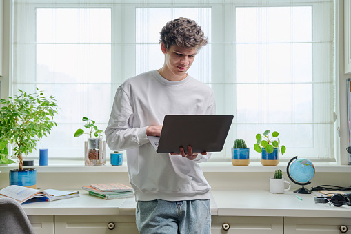 Portrait of friendly young college student guy holding laptop, at home interior. Handsome male 18-20 years old using laptop. Online internet technologies remote e-learning study leisure communication
