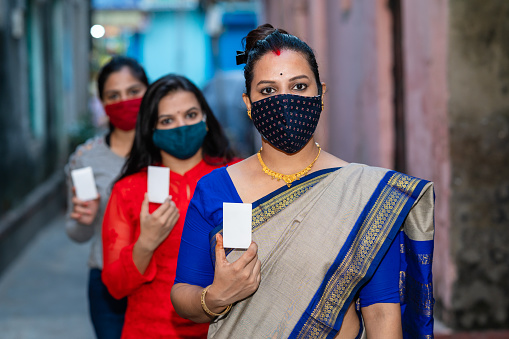 Outdoor image of Three young Women wearing face mask shows her voter id card or identity card while standing in a queue for casting their vote in Election.