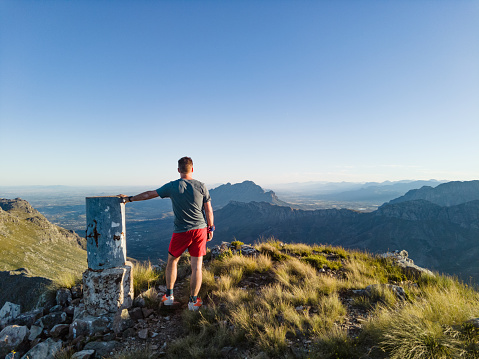 Rear view white mid-adult male trail runner standing at the cairn beacon on Haelkop Mountain Summit looking out over Jonkershoek, Stellenbosch, South Africa