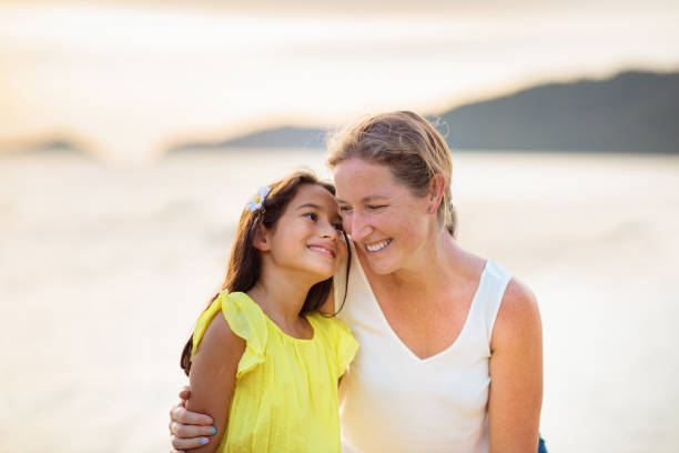 Mother and daughter on tropical beach. Summer sea vacation. Happy interracial family playing outdoor. Mom holding little girl. Young woman and cute child play and laugh. Holiday happiness and love.