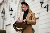 Beautiful young woman opening purse bag and taking something out while standing on street against of building. Stylish female model with fashionable hairstyle